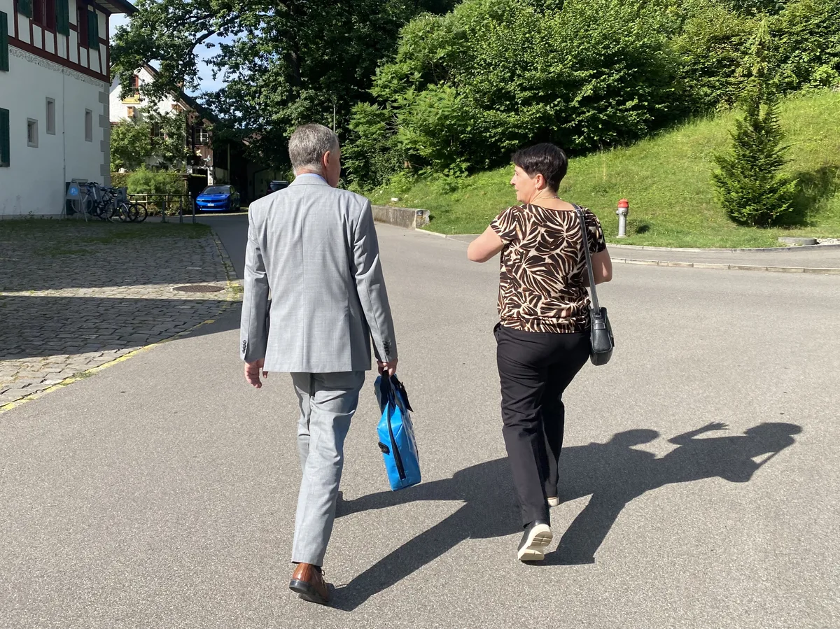 Eine Frau in gemusterter Bluse und ein Mann im hellgrauen Anzug von hinten.